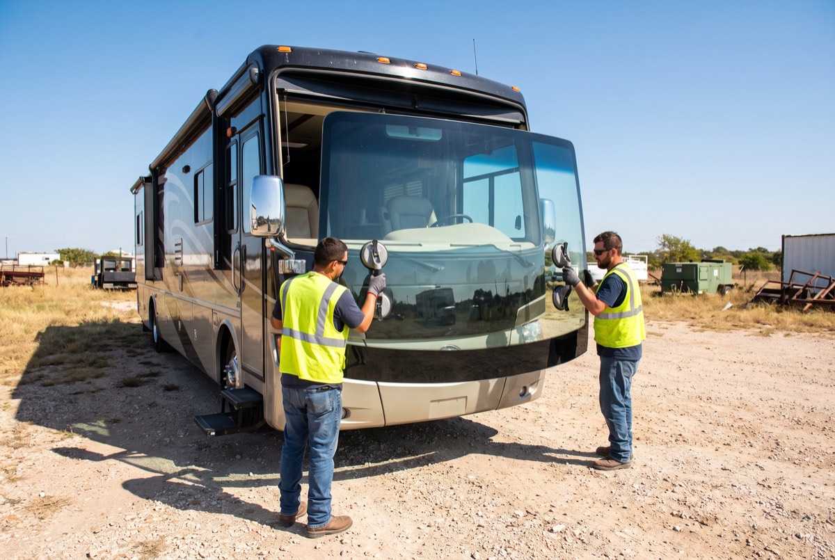 Two technicians setting a large Class A motorhome windshield with suction cups at a North Texas storage lot